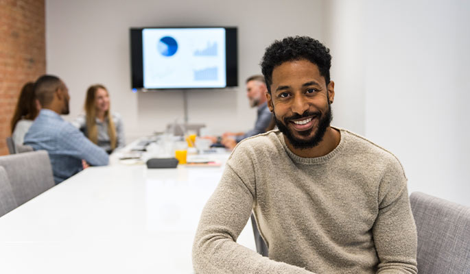 Bearded man seated at a conference table with a meeting in progress behind him